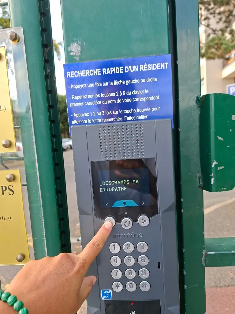 L’interphone du portail affichant « Deschamps MA étiopathe », un doigt sur le bouton de gauche.
