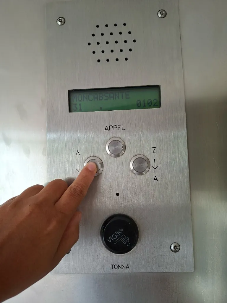 L’interphone du bâtiment affichant « MONCABSANTE », un doigt sur le bouton de gauche.